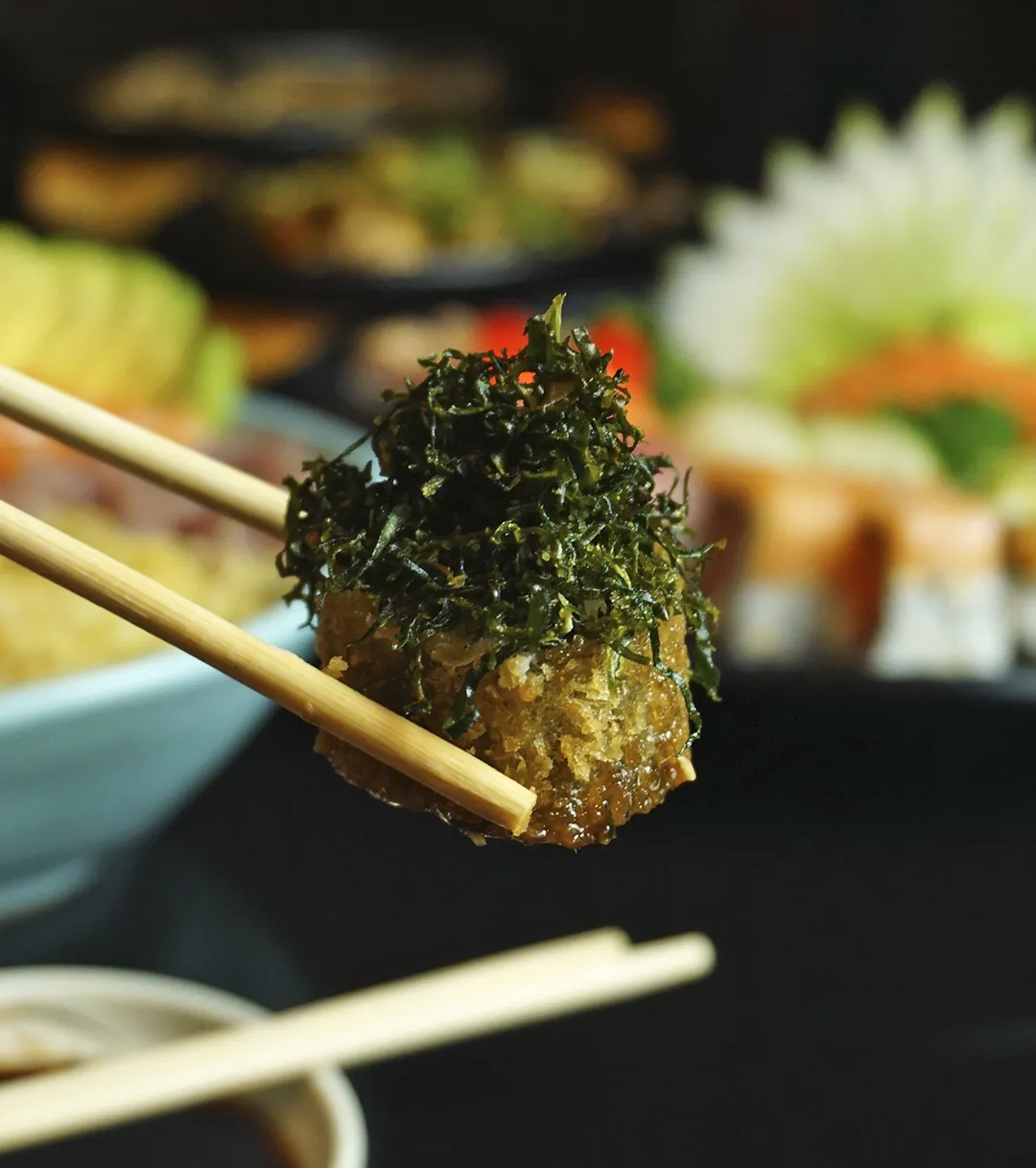 Pedaço de sushi frito coberto de alga, erguido com hashi sobre uma mesa de pratos variados
